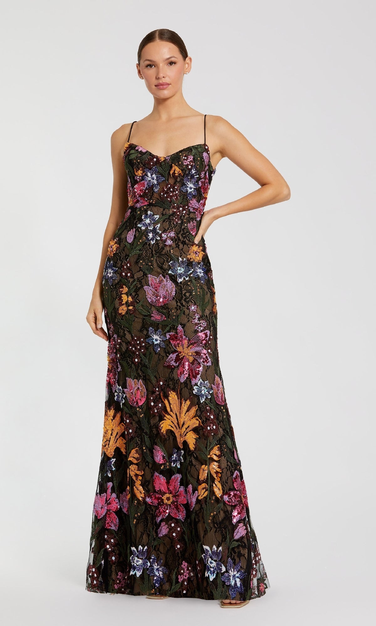 A woman wears the Mac Duggal 68885 Embroidered Long Formal Dressโa floor-length, sleeveless black gown with thin straps and a vibrant floral patternโstanding against a plain light background.